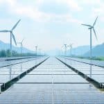 Solar panels are aligned in neat rows, with their shiny surfaces reflecting the sky. In the background, multiple wind turbines stand tall