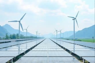 Solar panels are aligned in neat rows, with their shiny surfaces reflecting the sky. In the background, multiple wind turbines stand tall